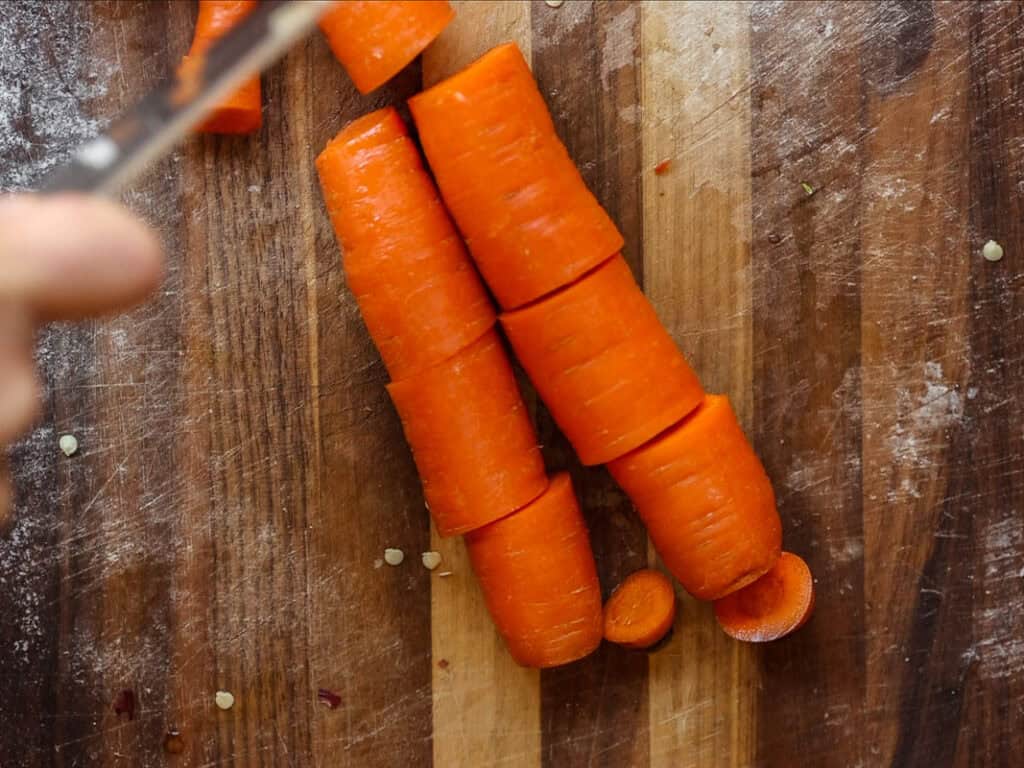 Close-up of chopped orange carrots on a wooden cutting board. A hand is holding a knife in the top left corner, mid-action. Small carrot pieces are scattered around.